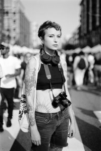An image of a non-binary person carrying a camera strapped around their neck. They are looking directly into the lens as a street festival is out of focus around them.